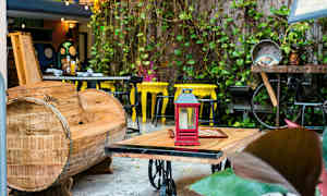 Ulkotila, jossa on puinen sohva, pöytä punaisella lyhdyllä ja keltaisia tuoleja taustalla. Kasvit koristavat ympäristöä. (Tekoälyn luoma teksti)