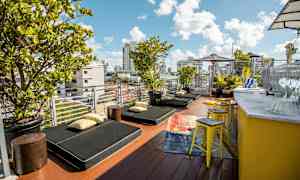 Terassi, jossa on aurinkotuoleja, tyynyjä ja vihreitä kasveja. Sivulla sijaitsee keltainen baaritiski jakkaroineen. (Tekoälyn luoma teksti)