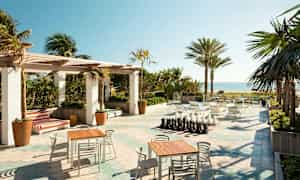 Ulkoterassi, jossa pöytiä ja tuoleja, shakkilauta sekä näkymä rannalle ja merelle, ympäröitynä palmuilla. (Tekoälyn luoma teksti)