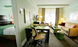 Hotellihuone, jossa vaalea sisustus, mukaan lukien sänky, kirjoituspöytä, sohva ja televisio. (Tekoälyn luoma teksti)