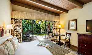 Hotellihuone, jossa on suuri sänky, pöytä ja tuolit sekä avoin liukuovi, joka johtaa vehreään ulkotilaan. (Tekoälyn luoma teksti)