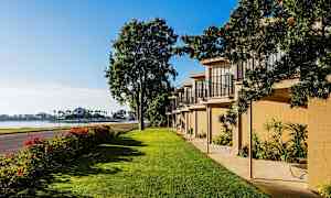 Kuvassa on vehreä alue, jossa on nurmikkoa, rivi rakennuksia parvekkeineen ja tie vieressä. Taustalla näkyy vettä ja palmuja. (Tekoälyn luoma teksti)