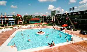 Ulkouima-allas, jonka ympärillä on aurinkotuoleja ja ihmisiä uimassa. Taustalla näkyy urheiluaiheisia rakennuksia, suuri koriste-esine, joka muistuttaa amerikkalaista jalkapalloa, sekä palmuja. (Tekoälyn luoma teksti)