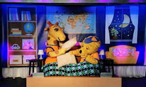 Kaksi piirrettyä hahmoeläintä, kirahvi ja karhu, istuvat sängyssä peiton kanssa, ja taustalla näkyy kohtauksen rekvisiittaa. (Tekoälyn luoma teksti)