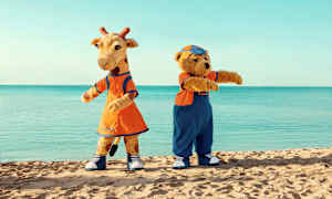 Kaksi maskottia, kirahvi ja karhu, seisovat rannalla taustanaan meri. (Tekoälyn luoma teksti)