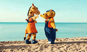 Kaksi maskottia, kirahvi ja karhu, seisovat hiekkarannalla taustalla näkyy meri. (Tekoälyn luoma teksti)