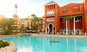 Hotellin lämminsävyinen julkisivu ja uima-allas sekä vihreää maisemaa. (Tekoälyn luoma teksti)