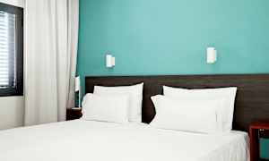 Hotellihuone, jossa on pedattu sänky valkoisilla lakanoilla ja sininen seinä. (Tekoälyn luoma teksti)