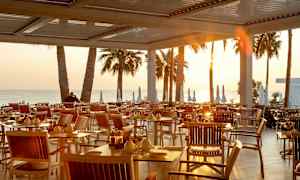 Ulkoravintolan pöydät ja tuolit auringonlaskussa, taustalla palmut ja meri. (Tekoälyn luoma teksti)