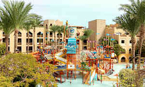 Kuva vesipuistosta, jossa on vesiliukumäkiä ja leikkirakenteita palmuineen ja hotellirakennuksien ympäröimänä. (Tekoälyn luoma teksti)