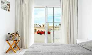 Valoisa makuuhuone, jossa on harmaa pedattu sänky, puutuoli ja sen päällä rullattu laukku, verhot sekä parvekkeen ovi, joka avautuu näkymään kukilla ja rakennuksilla. (Tekoälyn luoma teksti)