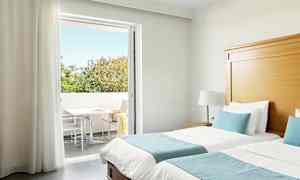 Hotellihuone, jossa on kaksi erillistä vuodetta, siniset vuodevaatteet ja suora pääsy parvekkeelle, jonka taustalla näkyy puita. (Tekoälyn luoma teksti)