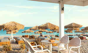 Näköala hiekkarannalle, jossa on aurinkovarjoja ja aurinkotuoleja, nähtynä istumapaikalta, jossa on pöytä ja tuoleja. (Tekoälyn luoma teksti)