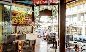 Kahvila-alue, jossa istuimia ja pöytiä ikkunan vieressä. Ulkona näkyy katu, jossa on liikkeitä ja autoja. (Tekoälyn luoma teksti)