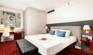 Hotellihuone, jossa suuri sänky, kaksi sinistä tyynyä, kirjoituspöytä tuoleineen, punainen matto ja nykyaikaiset valaisimet. (Tekoälyn luoma teksti)