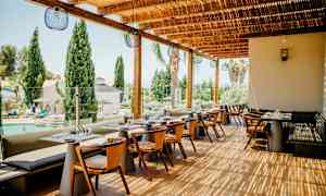 Ravintolan ulkoterassi, jossa pöytiä ja tuoleja, näkymä altaalle ja puille. (Tekoälyn luoma teksti)