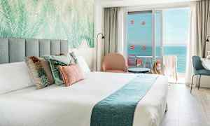 Hotellihuone, jossa on parivuode, koristeellisia tyynyjä, istuinryhmä ja parveke merinäköalalla. (Tekoälyn luoma teksti)