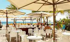 Ulkoilmaravintola, jossa valkoiset pöydät ja tuolit varjojen alla, näkymä merelle. (Tekoälyn luoma teksti)