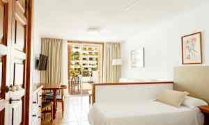 Hotellihuone, jossa on sänky, sohva ja ruokapöytä sekä parvekenäkymä, jossa on vehreyttä ulkona. (Tekoälyn luoma teksti)