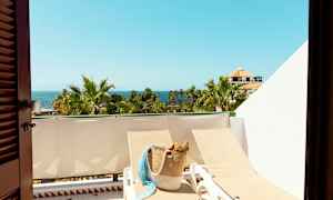 Näkymä aurinkoiselle parvekkeelle, jolla on aurinkotuoleja, korilaukku, palmuja ja meri taustalla. (Tekoälyn luoma teksti)