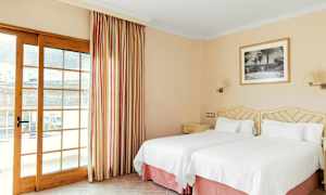 Hotellihuone, jossa kaksi yksittäistä vuodetta valkoisin vuodevaattein, seinällä taulu ja lasiovi verhoineen. (Tekoälyn luoma teksti)