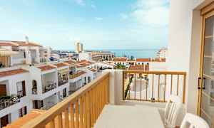 Näkymä parvekkeelta, jossa on pöytä ja tuolit, valkoisten rakennusten yli punaisine kattoineen ja taustalla meri. (Tekoälyn luoma teksti)