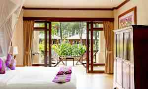 Makuuhuone, jossa sänky ja violetteja tyynyjä, näkymä puutarhaan suurten ikkunoiden ja ovien läpi. (Tekoälyn luoma teksti)