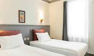 Kaksi sänkyä, joissa on valkoiset lakanat ja punaiset tyynyt, ovat huoneessa, jolla on vaaleat seinät ja seinälamppu. Oikealla on ikkuna valkoisilla verhoilla. (Tekoälyn luoma teksti)