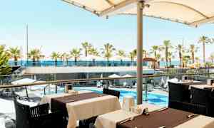 Näkymä ulkoilmaravintolasta kohti uima-allasaluetta, jossa on aurinkovarjoja ja palmuja sekä taustalla meri. (Tekoälyn luoma teksti)