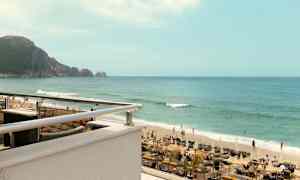 Kuva rannasta, jossa on aurinkotuoleja, aurinkovarjoja ja ihmisiä uimassa meressä. Taustalla näkyy kallioluoto. (Tekoälyn luoma teksti)