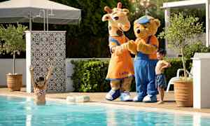 Kaksi hahmoa pukeutuneina kirahviksi ja nallekarhuksi seisovat altaan reunalla aurinkoisessa ympäristössä. (Tekoälyn luoma teksti)