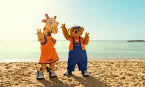 Kaksi maskotihahmoa nimeltään Lollo ja Bernie seisovat rannalla kirkkaan meren edessä. (Tekoälyn luoma teksti)