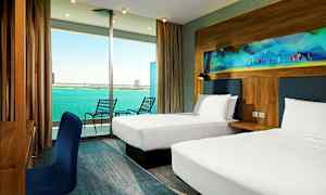 Hotellihuone, jossa on kaksi yhden hengen vuodetta, kirjoituspöytä sinisellä tuolilla ja parveke, josta on näkymä merelle. (Tekoälyn luoma teksti)