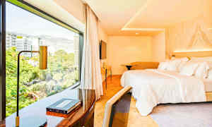 Hotellihuone, jossa on suuri sänky ja työpöytä lähellä suurta ikkunaa, joka näyttää näkymän yli kaupungin. (Tekoälyn luoma teksti)