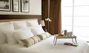 Huone, jossa on pedattu parisänky valkoisilla vuodevaatteilla ja aamiaistarjotin, jolla on croissantteja, kahvipannu ja kuppeja. Taustalla näkyy ruskeita verhoja ja tauluja seinällä. (Tekoälyn luoma teksti)