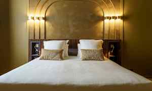 Hotellisänky valkoisilla vuodevaatteilla ja kultasävyisillä tyynyillä huoneessa, jossa on koristeellinen taustalevy. (Tekoälyn luoma teksti)