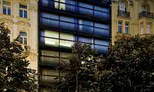 Kuvassa on moderni rakennus, jossa on tummat lasijulkisivut ja klassisia rakennuksia molemmin puolin, etualalla ravintola ja pysäköintialue. (Tekoälyn luoma teksti)