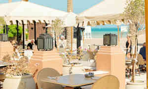 Ulkoterassi, jossa pöytiä ja tuoleja aurinkovarjojen alla lähellä rantaa. (Tekoälyn luoma teksti)