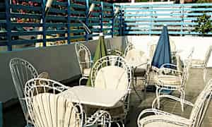 Terassi, jossa on valkoisia puutarhatuoleja ja pöytiä, siniseksi maalattu aitarakenne sekä kukkivat pensaat. (Tekoälyn luoma teksti)