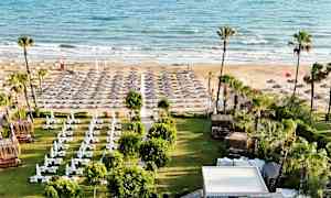 Kuva rannasta, jossa on aurinkotuoleja ja aurinkovarjoja sekä vihreä alue varjoa antavalla katolla edessä. (Tekoälyn luoma teksti)