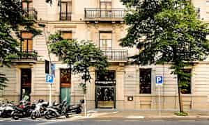 Rakennuksen kuva, jossa on vaaleasta kivestä tehty julkisivu. Rakennuksessa on useita parvekkeilla varustettuja kerroksia, ja sitä ympäröivät vihreät puut. (Tekoälyn luoma teksti)