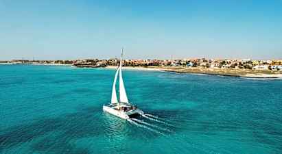 Katamaraani, jossa on valkoiset purjeet, kulkee turkoosissa vedessä lähellä rantaa, jolla on erivärisiä taloja. (Tekoälyn luoma teksti)