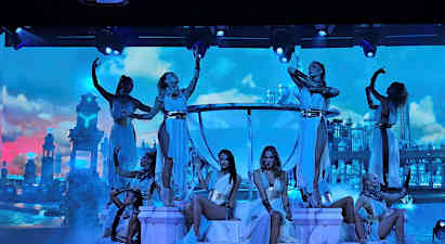 Tanssijat poseeraavat valaistulla näyttämöllä palatsimaisia rakennuksia sisältävän taustan edessä pilvien täyttämällä taivaalla. (Tekoälyn luoma teksti)