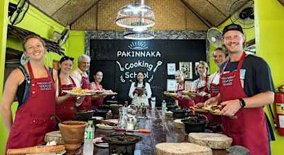 Ryhmää ihmisiä esiliinoissaan seisoo kyltin edessä, jossa lukee 'Pakinnaka Cooking School' keittiöstudiossa. (Tekoälyn luoma teksti)