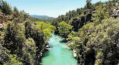 Turkoosin sininen joki virtaa vehreän rotkon läpi kallioiden ja puiden keskellä. (Tekoälyn luoma teksti)