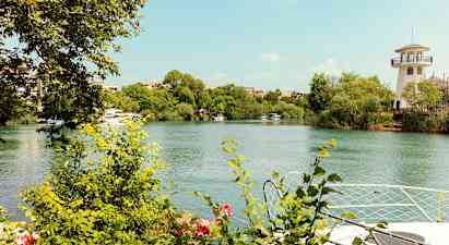 Joen rannat ovat täynnä vihreitä pensaita ja puita. Oikealla näkyy majakkaa muistuttava rakennus. (Tekoälyn luoma teksti)