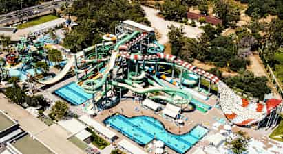 Vesipuisto, jossa on useita suuria vesiliukumäkiä, altaita ja aurinkotuoleja, ympäröitynä vihreillä puilla ja rakennuksilla. (Tekoälyn luoma teksti)