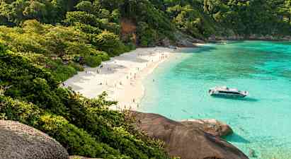 Hiekkaranta ihmisten kanssa, vehreiden puiden ympäröimä ja kirkkaan turkoosin veden reunustama. Vene on lähellä rantaa. (Tekoälyn luoma teksti)