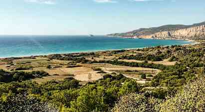 Maisemakuva, jossa vihreitä peltotiloja, pensaita ja vuoria ulottuu rannalle, turkoosi meri kirkkaan taivaan alla. (Tekoälyn luoma teksti)