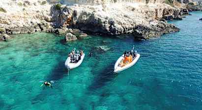 Kaksi venettä ankkuroituina kirkkaassa turkoosissa vedessä lähellä kallioista rantaa. Sukeltajat ovat vedessä. (Tekoälyn luoma teksti)